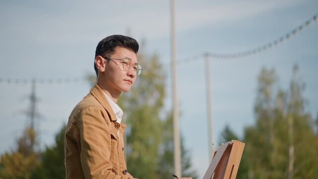 outdoor artist painting on easel, man in brown jacket and glasses focuses on sketchbox in sunny park with string lights and distant power lines, wooden palette and brushes, loose brushstrokes, calm