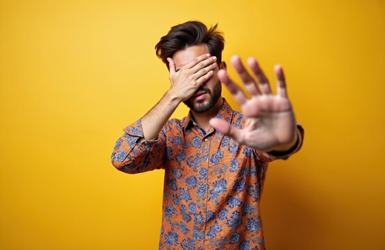 Man with hand over eyes makes stop sign. He hides face, looking away from camera with palm out. Person feels shy or scared, wants privacy. Isolated on yellow.