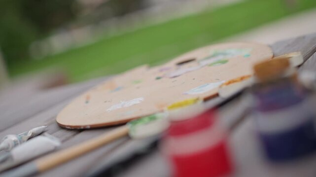 closeup palette on wooden bench paints jars brushes blurred background, hand testing pigment from tube, fingertips smearing dab of color, casual jacket sleeve, park greenery and midday sunlight,