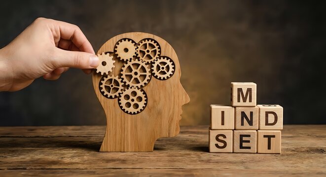 Hand placing gear into wooden head silhouette with brain made of cogs next to mindset letter blocks on dark background representing mental development.