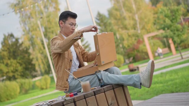 asian artist sketching on park bench, mixing watercolor and opening sketchbook, arranging paints on wooden seat, casual jacket and sneakers, leafy trees and paved path in background, relaxed urban