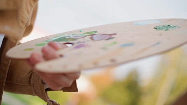 closeup palette mixing paint by white, artist holding wooden palette dabs blending sky blue and green pigments, jacket sleeve visible, sunlit park backdrop, textured brush tips, careful color