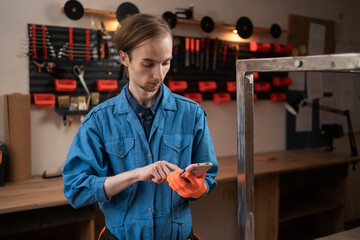 Young craftsman in overalls using smartphone for online shopping in workshop