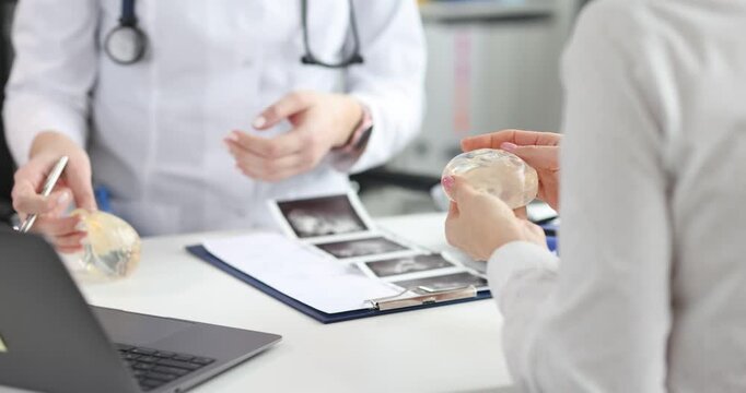 Woman doctor holds breast implant sample explaining augmentation result. Female patient tests silicone implant in hand imagining chest shape