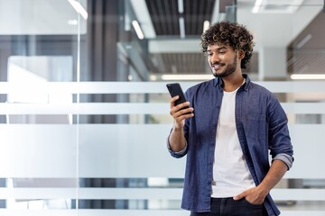Indian young smiling man standing in office, holding hand in pocket and using mobile phone