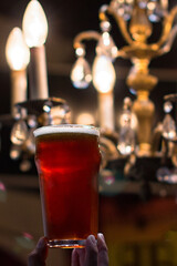 Glass of red amber craft beer held up against elegant chandelier lights in a dark bar interior. Warm evening atmosphere, premium pub concept.