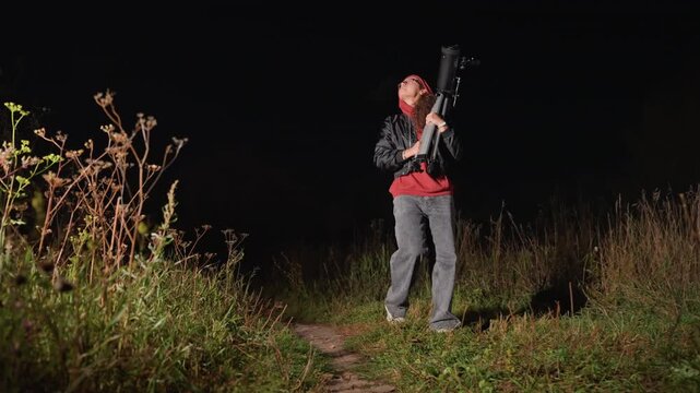 Night sky stargazing with telescope, outdoors person in red hoodie adjusts tripodmounted refractor on grassy meadow path, focused on distant stars, quiet atmosphere with soft flashlight glow,