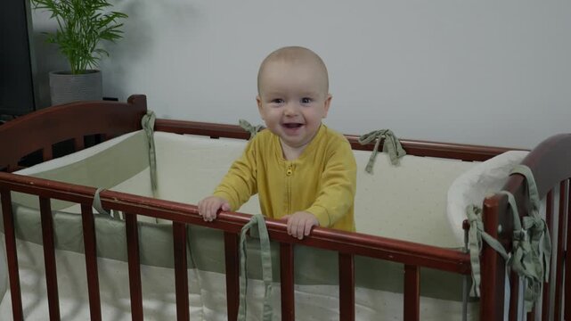caucasian baby standing at crib smiling broadly, milestone achiever in yellow onesie showing joyful grin and playful gaze, bright nursery with potted plant and modern decor capturing