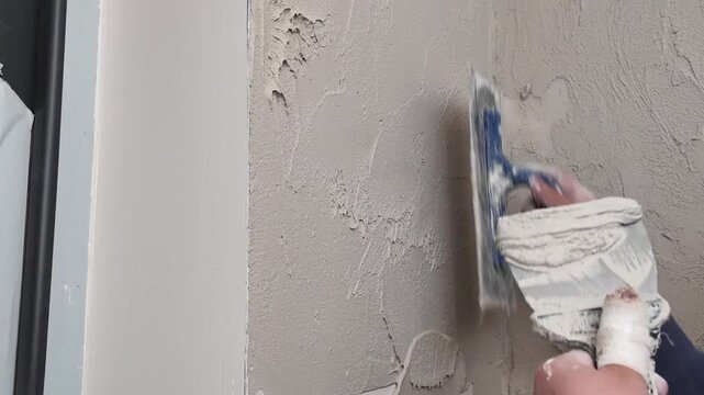 Hands are seen applying plaster to a wall during a home improvement project in a living room. The process involves smoothing and finishing the surface for renovation.