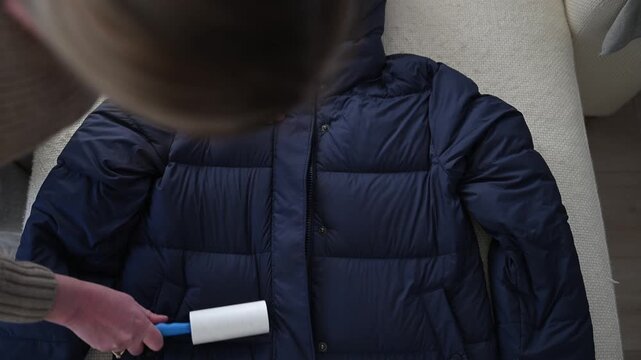 Woman removing pet hair from winter jacket using lint roller at home, fabric care concept