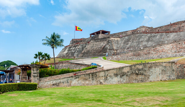 The massive stone walls of San Felipe de Barajas Castle rise above green lawns and palm trees under a cloudy sky in the coastal city of Cartagena, Bol&iacute;var, Colombia