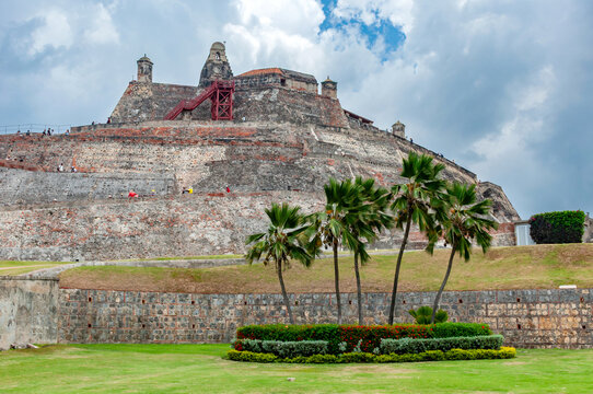 The massive stone walls of San Felipe de Barajas Castle rise above green lawns and palm trees under a cloudy sky in the coastal city of Cartagena, Bol&iacute;var, Colombia