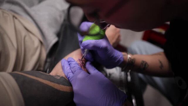 Close up shot of a professional tattoo artist wearing purple gloves and making a permanent tattoo on a client's arm with an electric needle machine in a modern studio