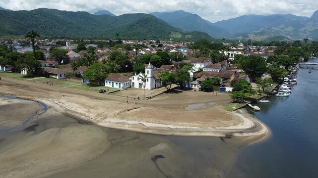Drone flying from the bay of Paraty towards the town and beach by the river, with the mountains and the Atlantic forest that surrounds this historical city