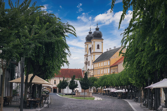 Historic square in Trenč&iacute;n, Slovakia&mdash;Baroque church with onion domes, tree-lined plaza, colorful townhouses, and outdoor caf&eacute;s under a blue summer sky.
