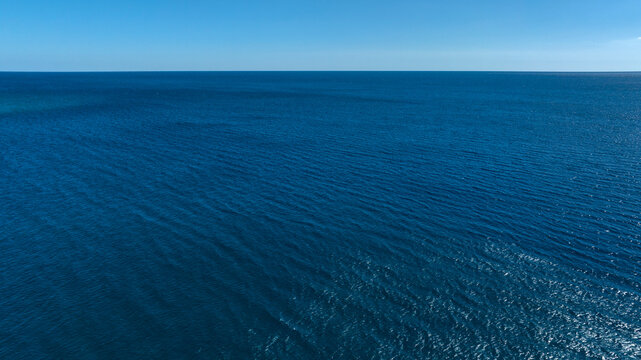 A vast, high-angle view of a deep blue sea with gentle ripples on the surface, meeting a clear, light blue horizon under a bright sky.
