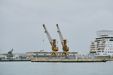 Obraz premium Two yellow harbor cranes standing idle on a concrete quay beside a large white cruise ship under grey sky