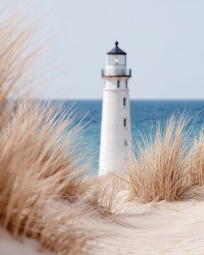 Lighthouse standing tall amidst coastal grasses on sandy dunes, with a clear blue sky and ocean in the background, creating a serene minimalist scene