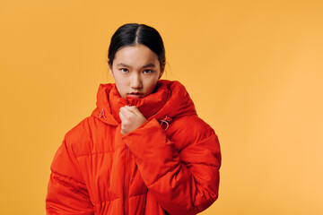 Young woman in red puffer jacket with high collar covering mouth, standing against orange...
