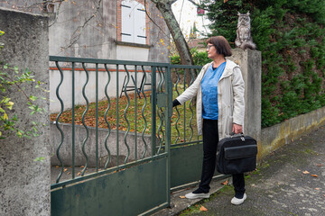 A woman stands at a gate with a bag while a cat watches from a wall