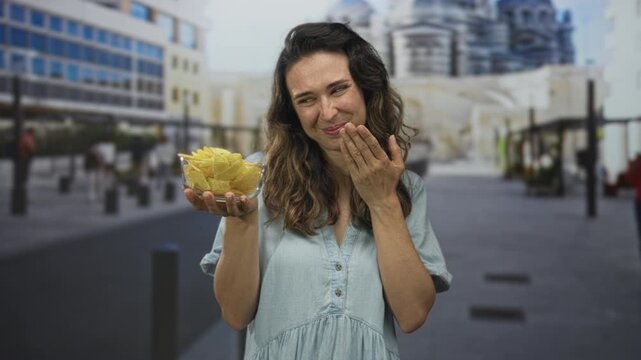 Woman smiling and holding glass bowl of crispy chips with hand to mouth gesture on street; happiness.