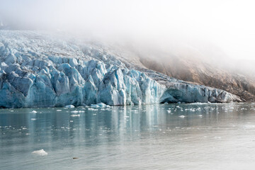 Close-up view of a massive glacier terminus meeting the ocean in East Greenland. Thick fog and mist shroud the upper ice and rocky cliffs, highlighting the deep blue crevasses.