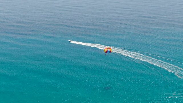 High angle aerial drone shot of speed boat pulling colorful parasail parachute over calm turquoise ocean water surface in summer