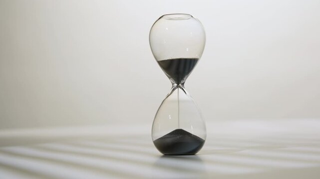 Side view of hourglass with black sand evenly flowing from one glass bulb to another on white desk close-up
