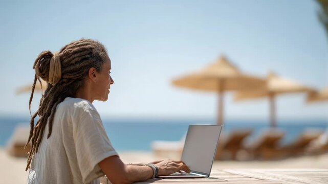 Young woman with dreadlocks, possibly a digital nomad or remote worker, sits at a wooden table on a sunny beach, typing on her laptop with the ocean and beach umbrellas in the background