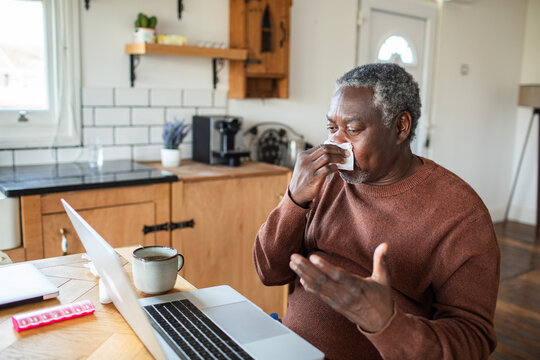 Older man using tissue during video call at home kitchen