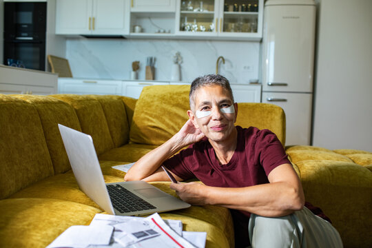 Mature woman paying bills online at home with under-eye patches