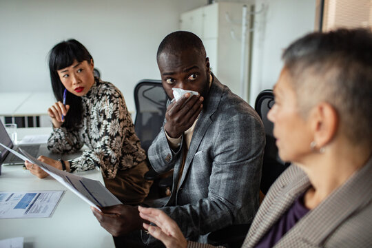 Coworkers in office meeting as sick colleague covers cough
