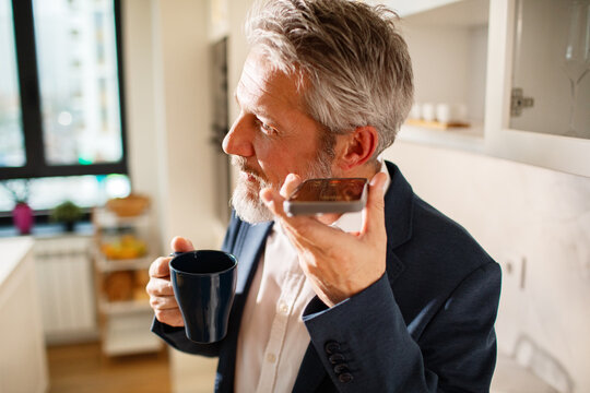 Mature businessman using smartphone speakerphone in home kitchen