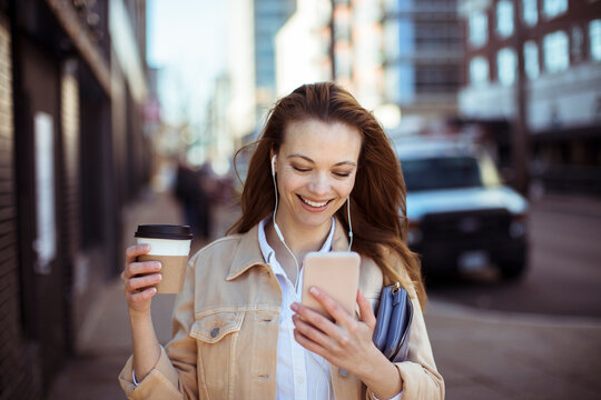 Smiling young woman with coffee using smartphone on city street