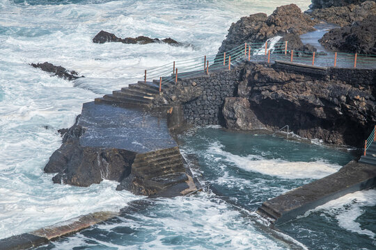 Escaleras de piedra y senderos de hormig&radic;&ge;n en una piscina natural de la costa, con olas blancas rompiendo contra las rocas volc&radic;&deg;nicas negras bajo la luz del d&radic;&ne;a