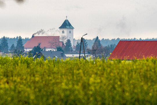 Wiosenny krajobraz wiejski z polem kwitnącego rzepaku na pierwszym planie oraz kościołem i zabudową z czerwonymi dachami w tle.