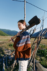 Obraz premium Woman axe wood countryside mountains sweater outdoor rural profile carrying large splitting axe over shoulder near wooden fence and stacked logs in mountain meadow landscape.