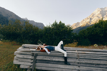 Obraz premium Woman resting on stacked wood beams in an orchard, surrounded by trees and mountains, outdoors relaxation scene with casual denim and boots, peaceful summer landscape.