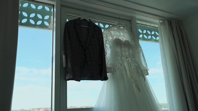 A wedding jacket and a wedding dress are hanging on the window