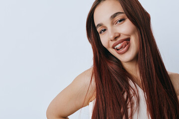 Obraz premium Young woman with long brown hair smiling playfully and sticking out her tongue against a plain light background. Casual portrait with natural expression and relaxed pose.