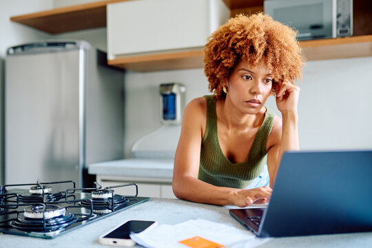 Woman focused at kitchen counter, working on laptop and managing finances with bills, credit card and phone in natural light