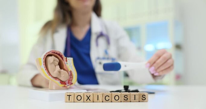 Text Toxicosis on wooden cubes and human fetus anatomical model in hospital. Female gynecologist shows positive pregnancy test in medical office