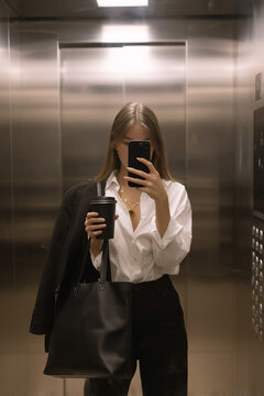 Junge Frau macht ein Spiegel-Selfie im Aufzug, elegant gekleidet mit Tasche in der Hand. Moderne Lifestyle-Fotografie mit urbaner Atmosph&auml;re. Ideal f&uuml;r Business, Karriere, Social Media, Alltag und zei