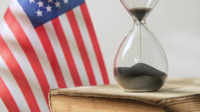 Hourglass on old codex book with American flag, sand pouring from one container to another, countdown, time limit for making decision