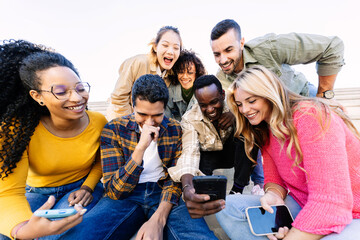 Fototapeta premium Multiracial group of young students enjoying funny content on a smartphone while relaxing together outdoors. Youth community and friendship lifestyle.