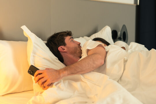 Unfaithful boyfriend texting on his mobile phone in bed while his brunette girlfriend sleeps peacefully next to him, capturing betrayal, hidden communication, lack of trust, and a secret affair