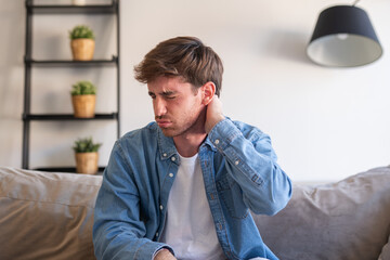 Fototapeta premium An upset and tired young man sits at home massaging his stiff neck and rubbing tense muscles, trying to relieve back and shoulder pain caused by long sedentary work and poor posture daily