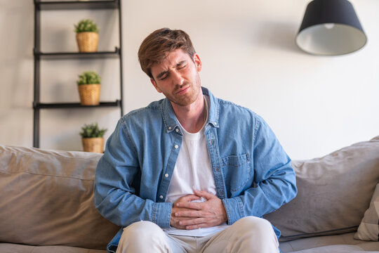 A sick young man sits at home holding his stomach in pain, suffering from abdominal discomfort caused by indigestion, gastritis, or digestive problems, showing symptoms like bloating, cramps, diarrhea