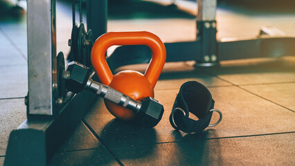 Orange kettlebell, black dumbbell, and ankle strap placed on gym floor, emphasizing strength training and fitness preparation.
