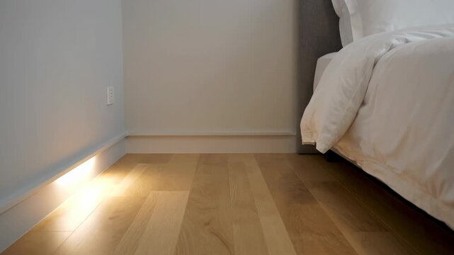 Subtle accent lighting illuminates a modern bedroom floor. Clean white walls complement the warm wooden planks.